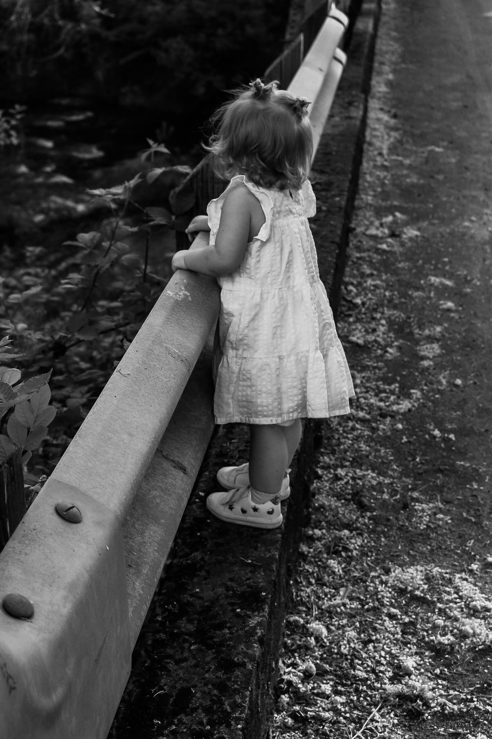 Black and white photo of a toddler girl in a white dress leaning over a bridge railing.