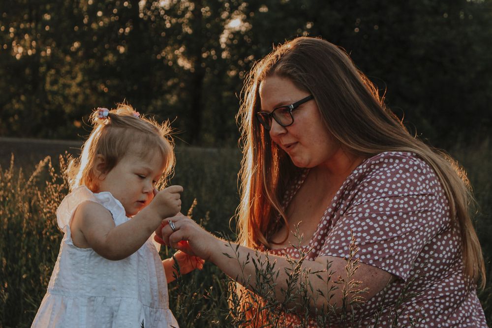 A mother with a tattoo helps her toddler daughter examine a flower during a warm sunset outdoor session.