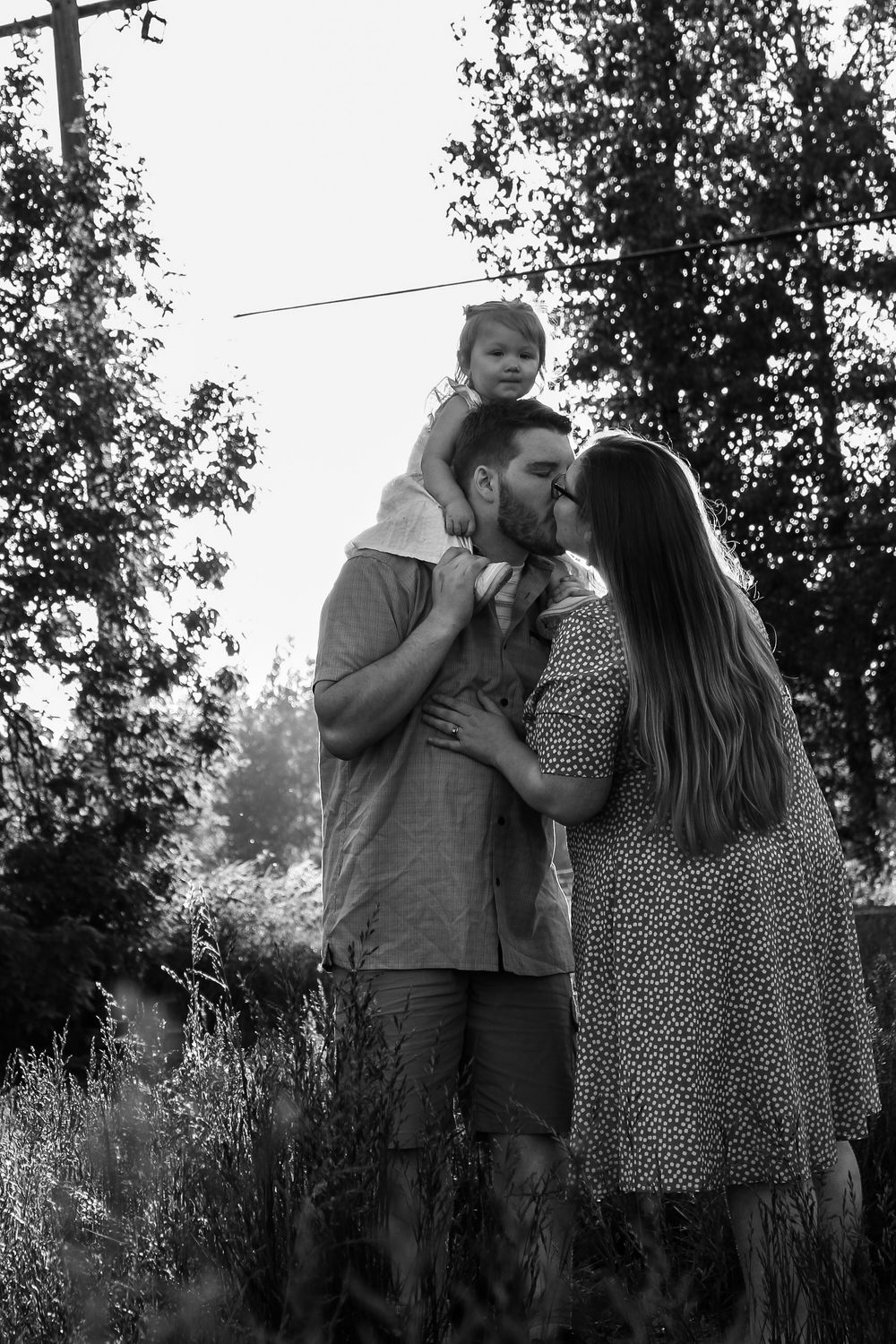 A couple kisses outdoors while their young child sits on the mans shoulders in a black and white family photo.
