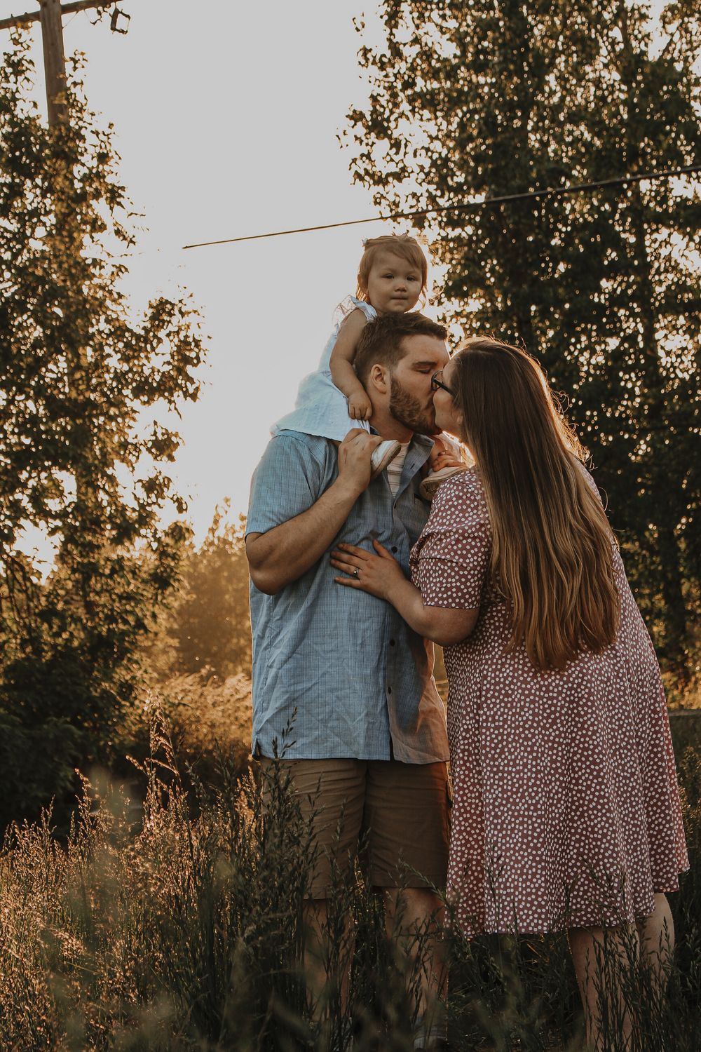 A couple shares a kiss while their toddler sits on the fathers shoulders during a golden hour family session.