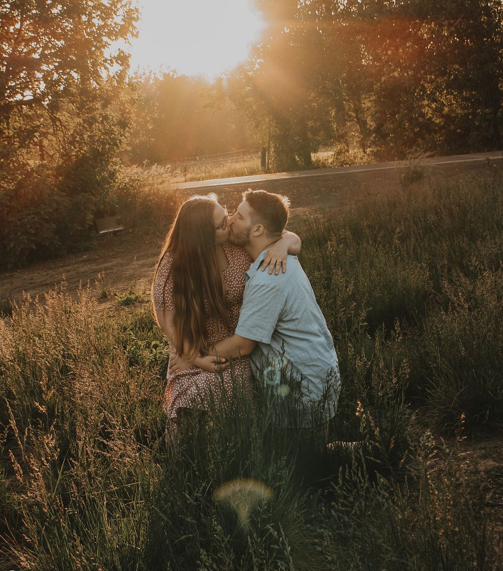 A couple shares a romantic kiss while sitting in a field of tall grass during a beautiful golden hour sunset.