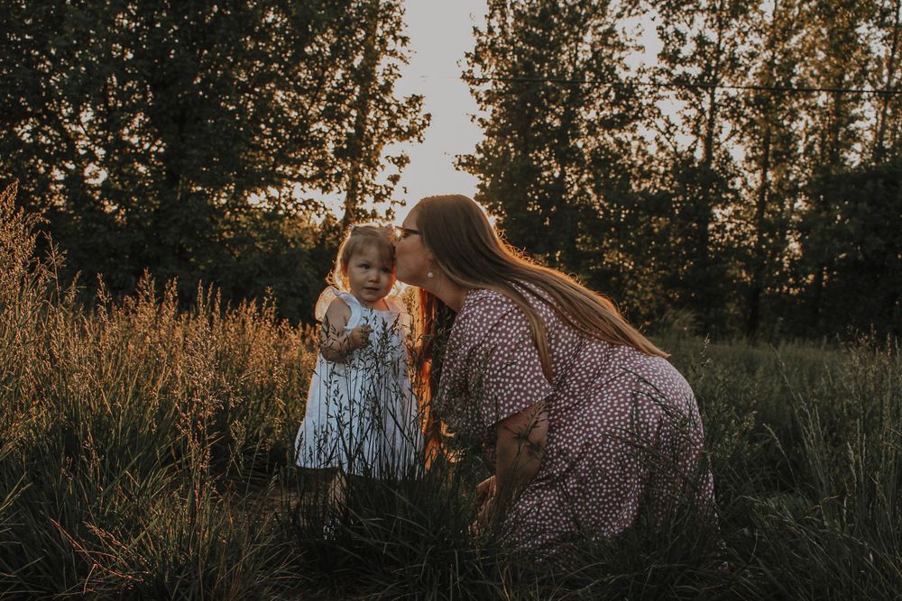 A mother kneels to kiss her toddler daughters cheek while surrounded by tall grass in golden evening light.