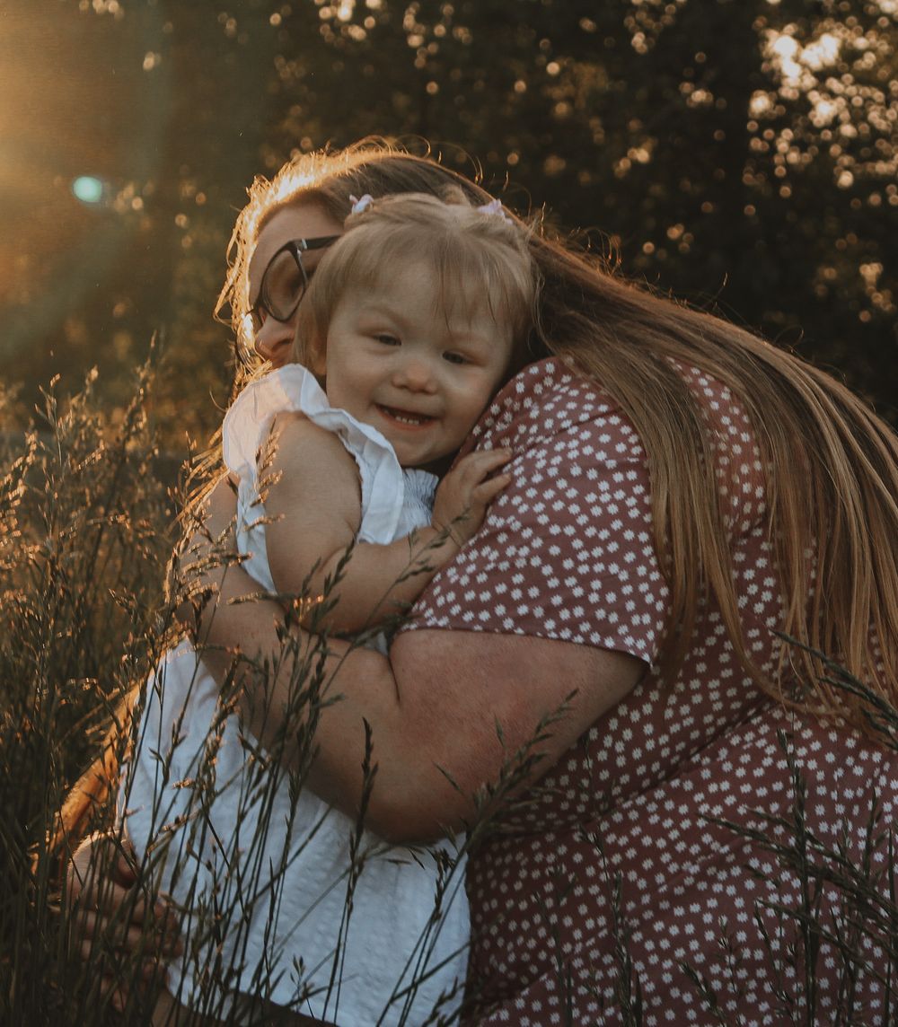 A mother warmly embraces her smiling toddler daughter in a sun-drenched field during a golden hour session.