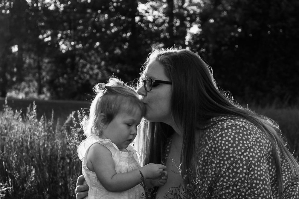 A mother wearing glasses kisses her toddler daughter on the cheek in a black and white outdoor portrait.