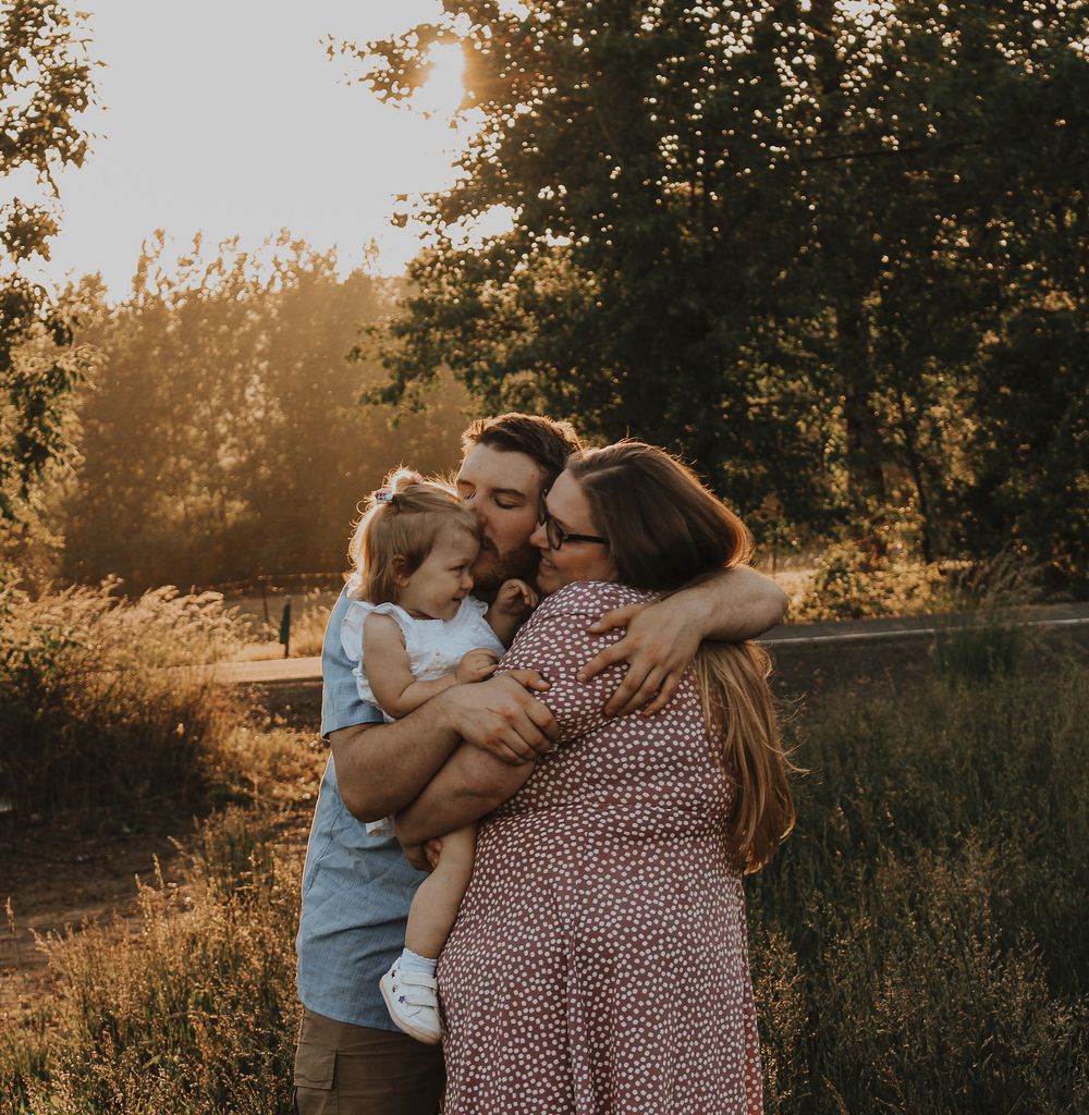 A family of three shares a warm embrace in a sunlit outdoor setting during a golden hour photo session.