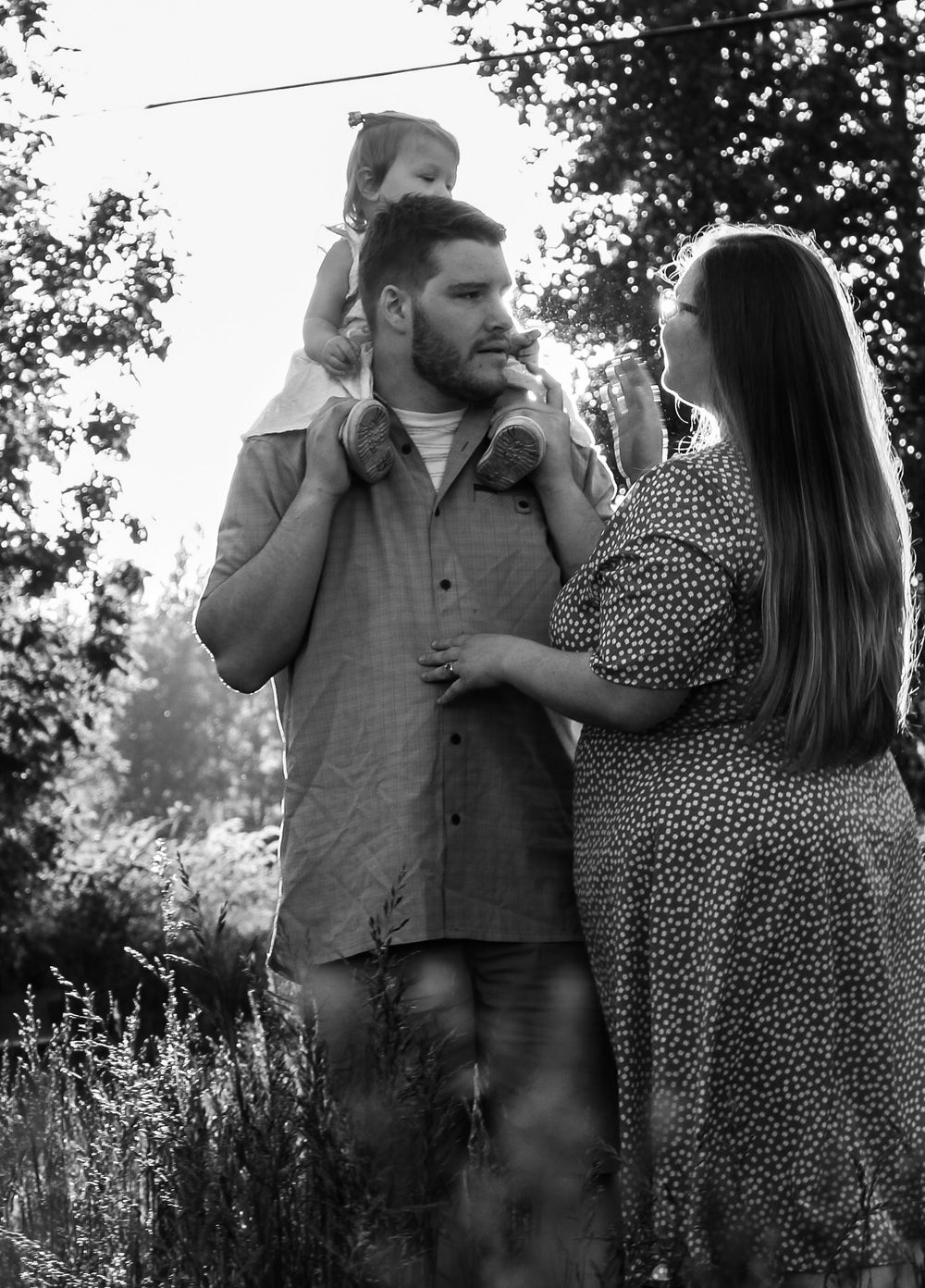 A man carries a toddler on his shoulders while a woman in a floral dress looks on in a black and white outdoor photo.