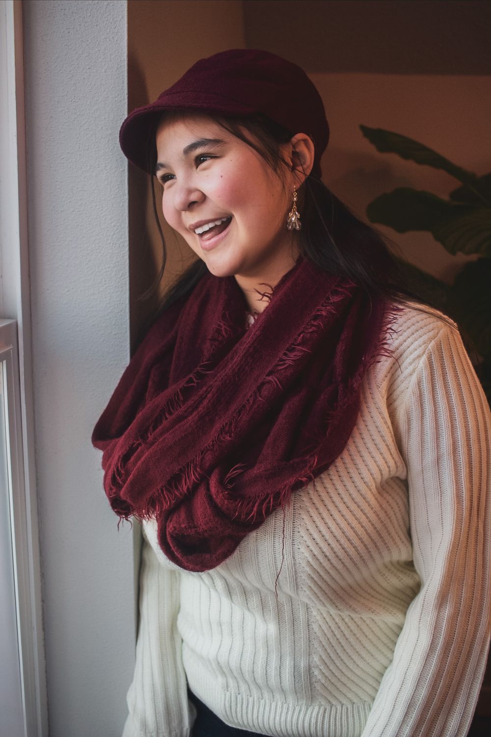 Smiling young woman wearing a burgundy cap and matching infinity scarf with a cream knit sweater.
