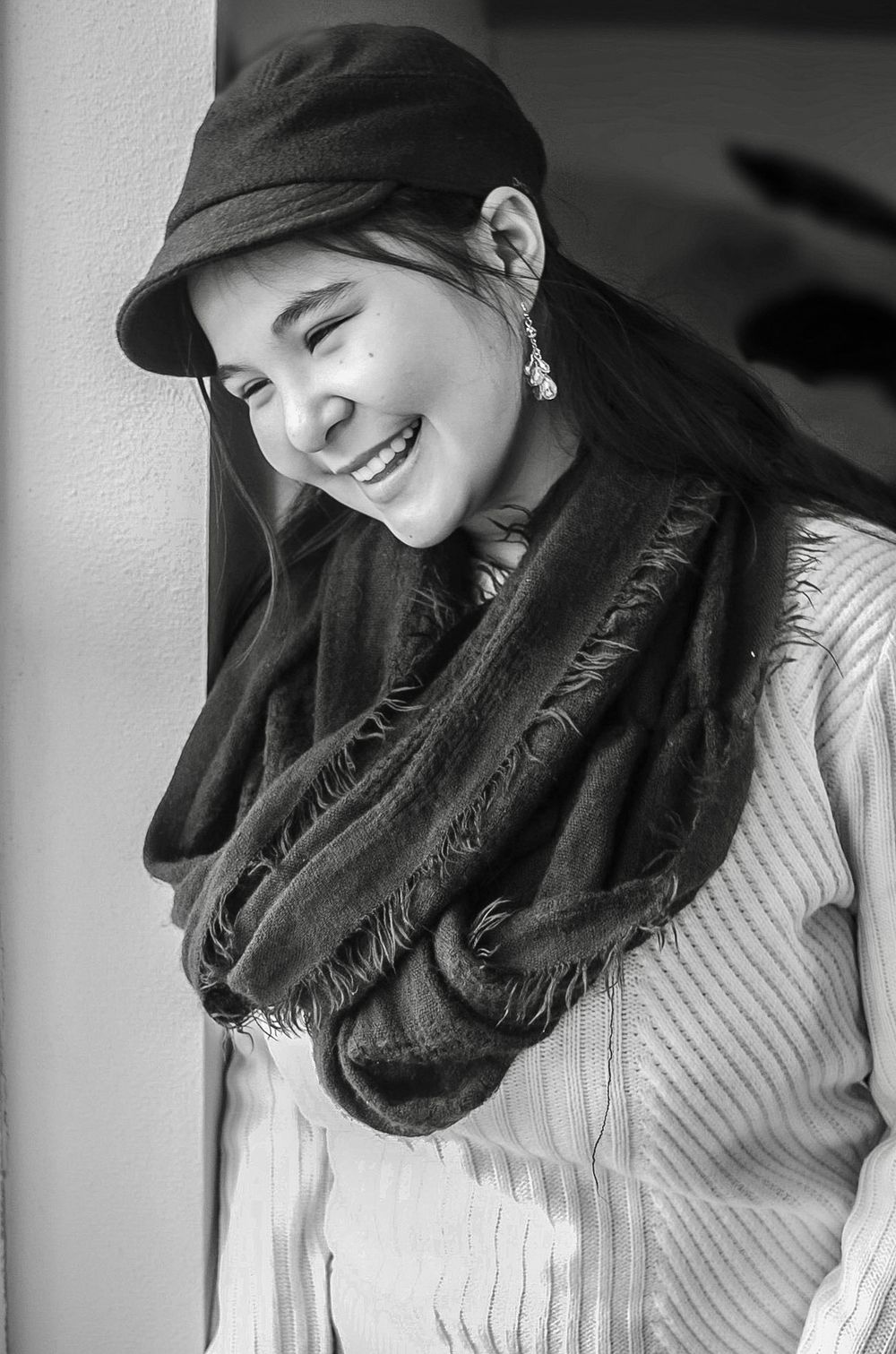Black and white portrait of smiling young woman in cap and scarf leaning against wall.