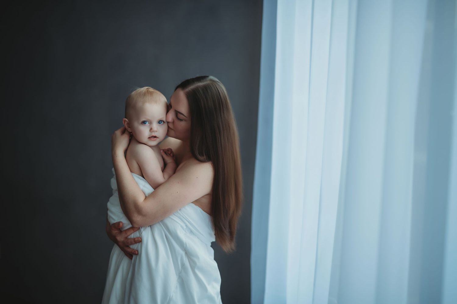 Mother wearing white dress gently embraces baby near window with soft window light and dark background.