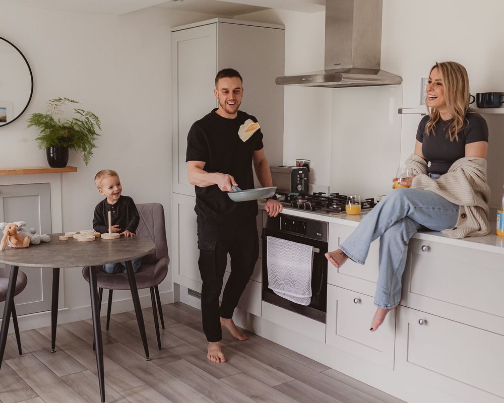 A man flips pancakes in a modern white kitchen while a woman sits on the counter and a toddler watches.
