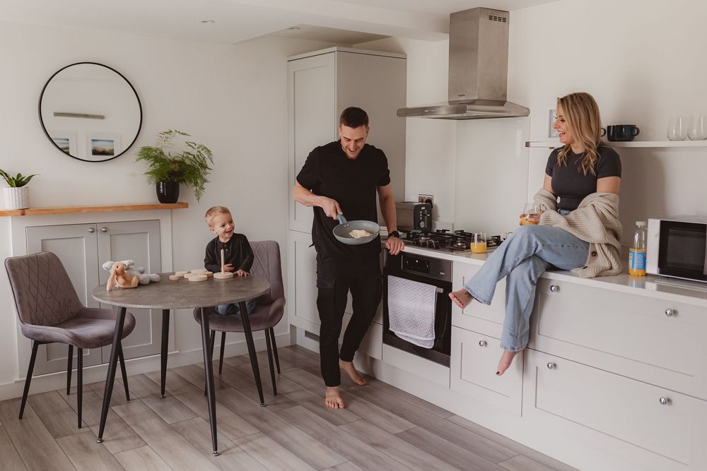 A young family enjoys a relaxed morning in a bright modern kitchen with a toddler seated at the table.