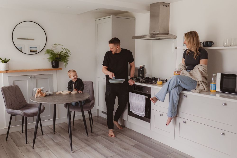 A couple and their toddler share a casual morning moment in a stylish white kitchen with modern appliances.