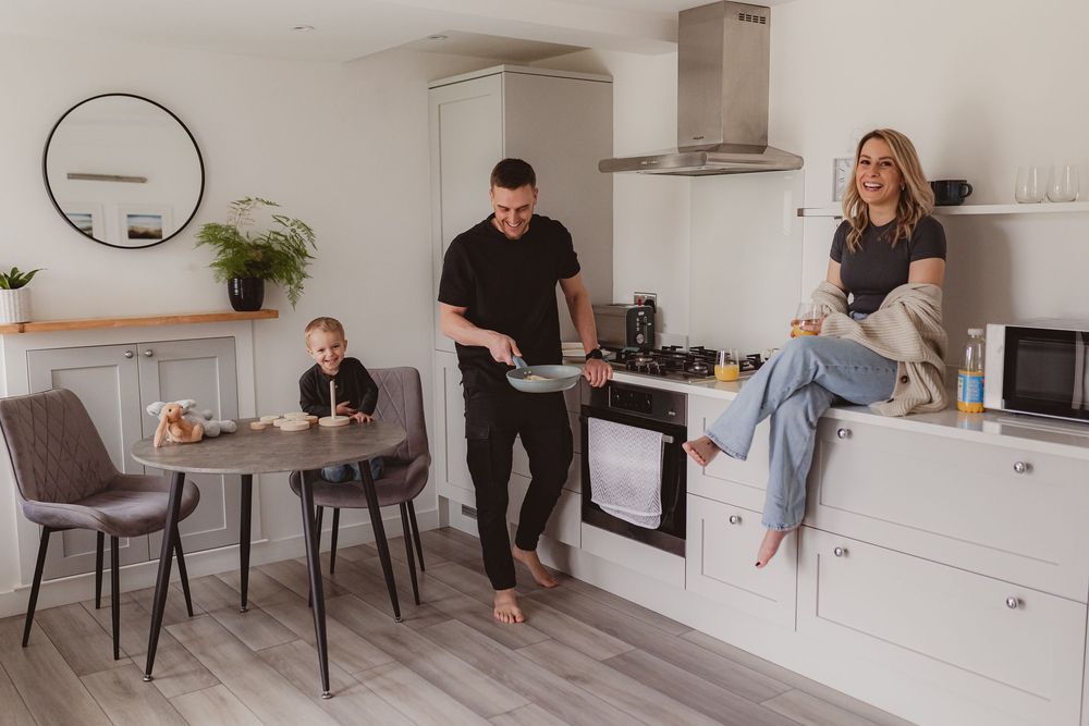 Family in modern white kitchen, man cooking while woman sits on counter and child plays at dining table.