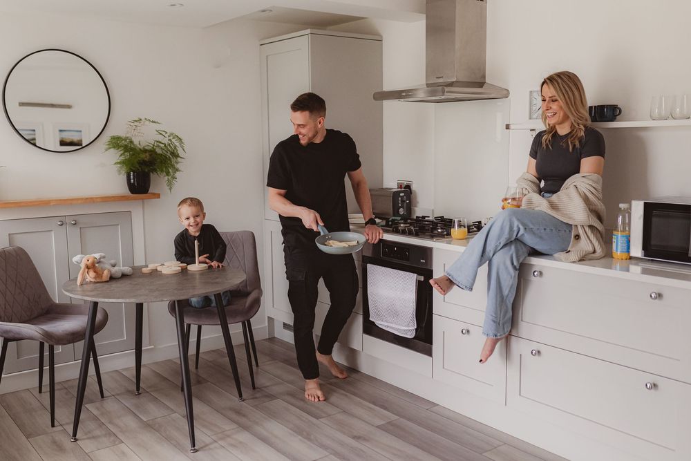 Young family enjoying morning in bright kitchen, dad cooking pancakes while mom relaxes on counter near child.
