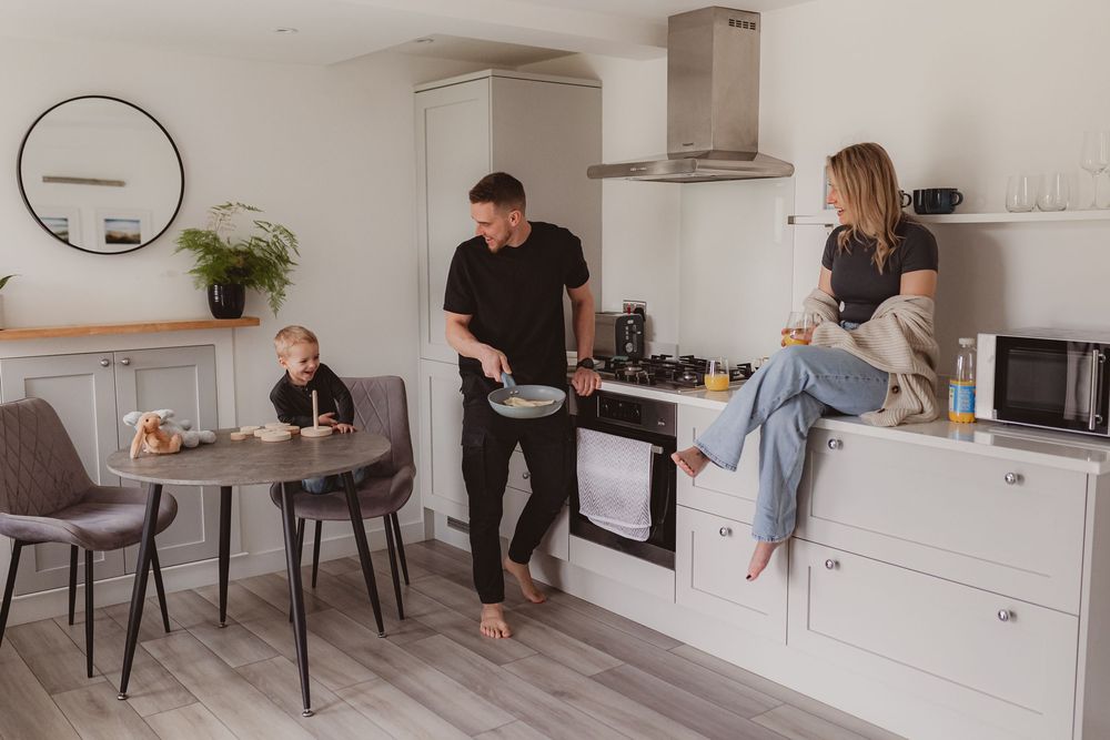 Happy family in contemporary white kitchen with dad cooking, mom sitting on counter, and toddler at table.