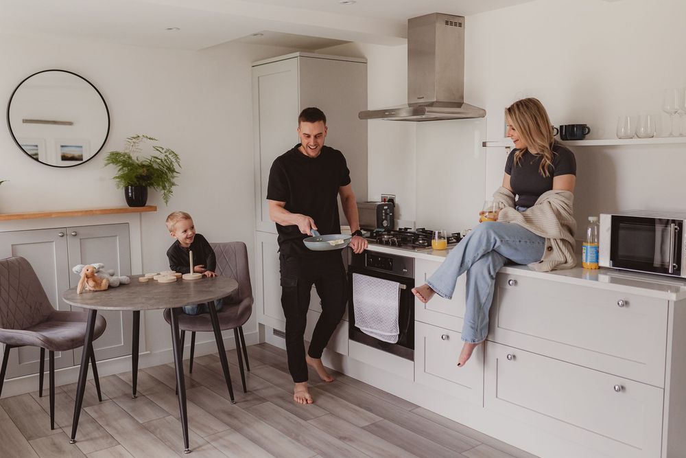 Family lifestyle photo in modern kitchen with man flipping pancakes, woman on counter, child at dining table.