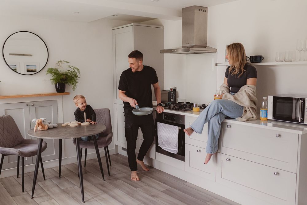 Smiling family in bright white kitchen, father cooking while mother sits on countertop and child plays nearby.