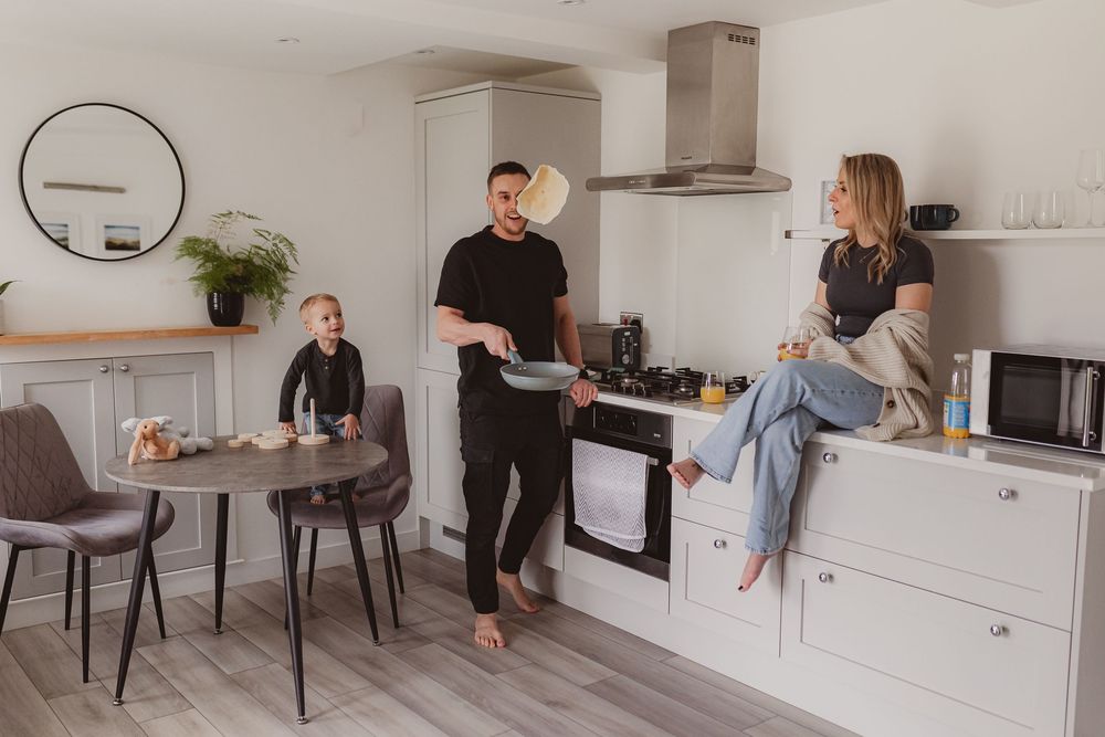 Dad flipping pancake in stylish white kitchen while mom watches from counter and toddler sits at table.