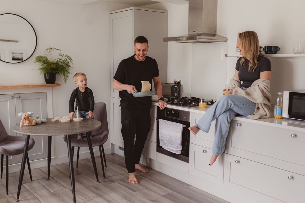 Family morning routine in modern kitchen with father cooking, mother relaxing on counter, child at round table.