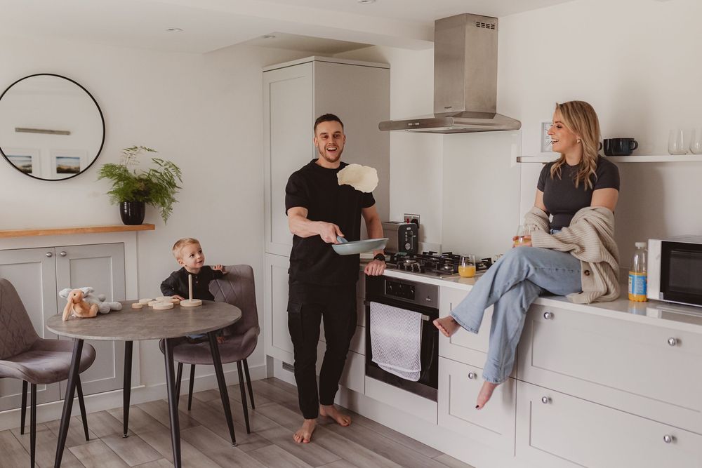 Father tossing pancake in white kitchen while laughing with mother on counter and child watches at table.
