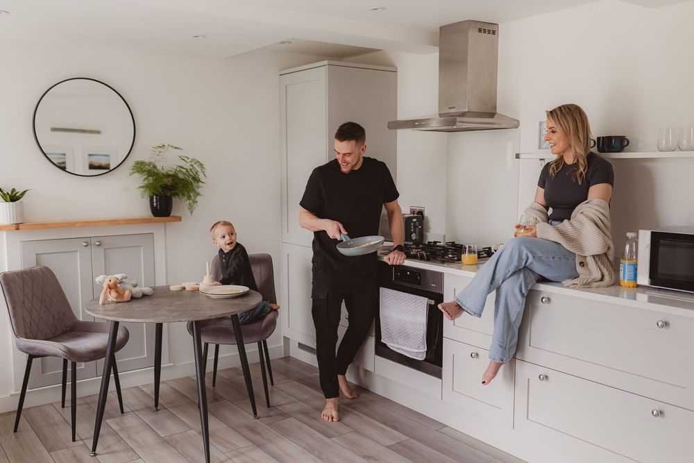 Contemporary white kitchen with family, dad cooking at stove, mom seated on counter, toddler at dining table.