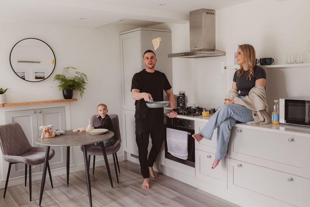 Family cooking pancakes in modern white kitchen, man flipping pancake while woman sits on counter and child plays at table.