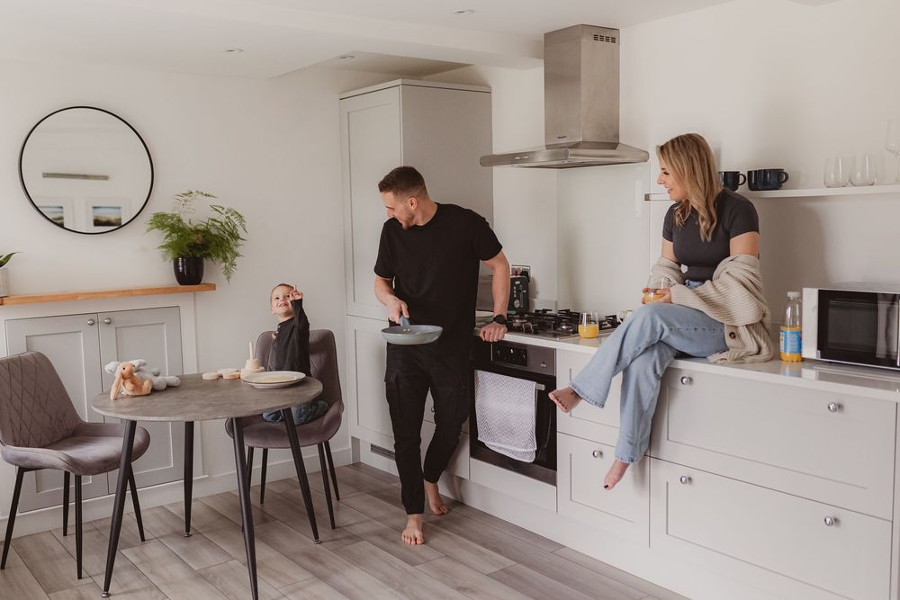 Happy family in bright white kitchen, man serving food while woman sits on counter and child sits at dining table.