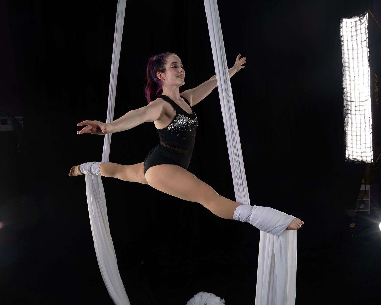 Aerial performer in black leotard executes splits pose on white silk ribbons against dark background.