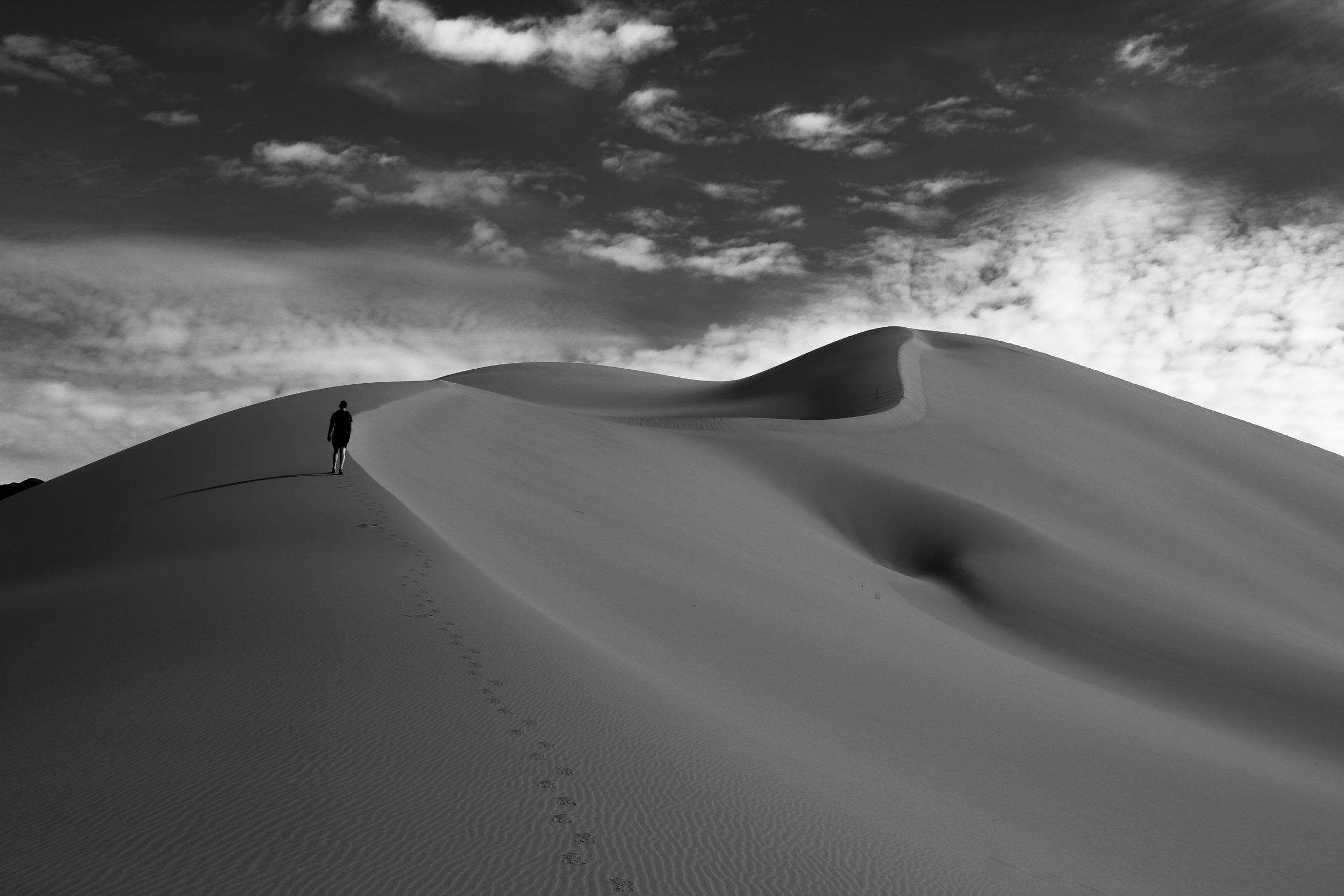 A black and white photo of a man hiking up the side of a sand dune. Photographed at Ibex Dunes in Death Valley National Park.