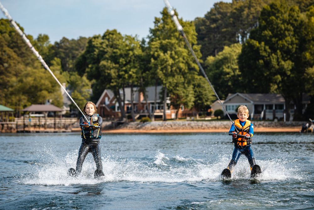 Two children water skiing side by side on a lake, wearing life jackets, with trees and buildings in the background.
