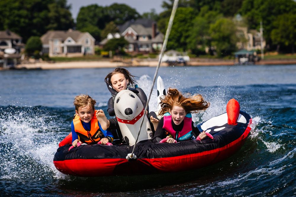 Three children riding a Snoopy-themed inflatable tube being towed across a lake, wearing life jackets.