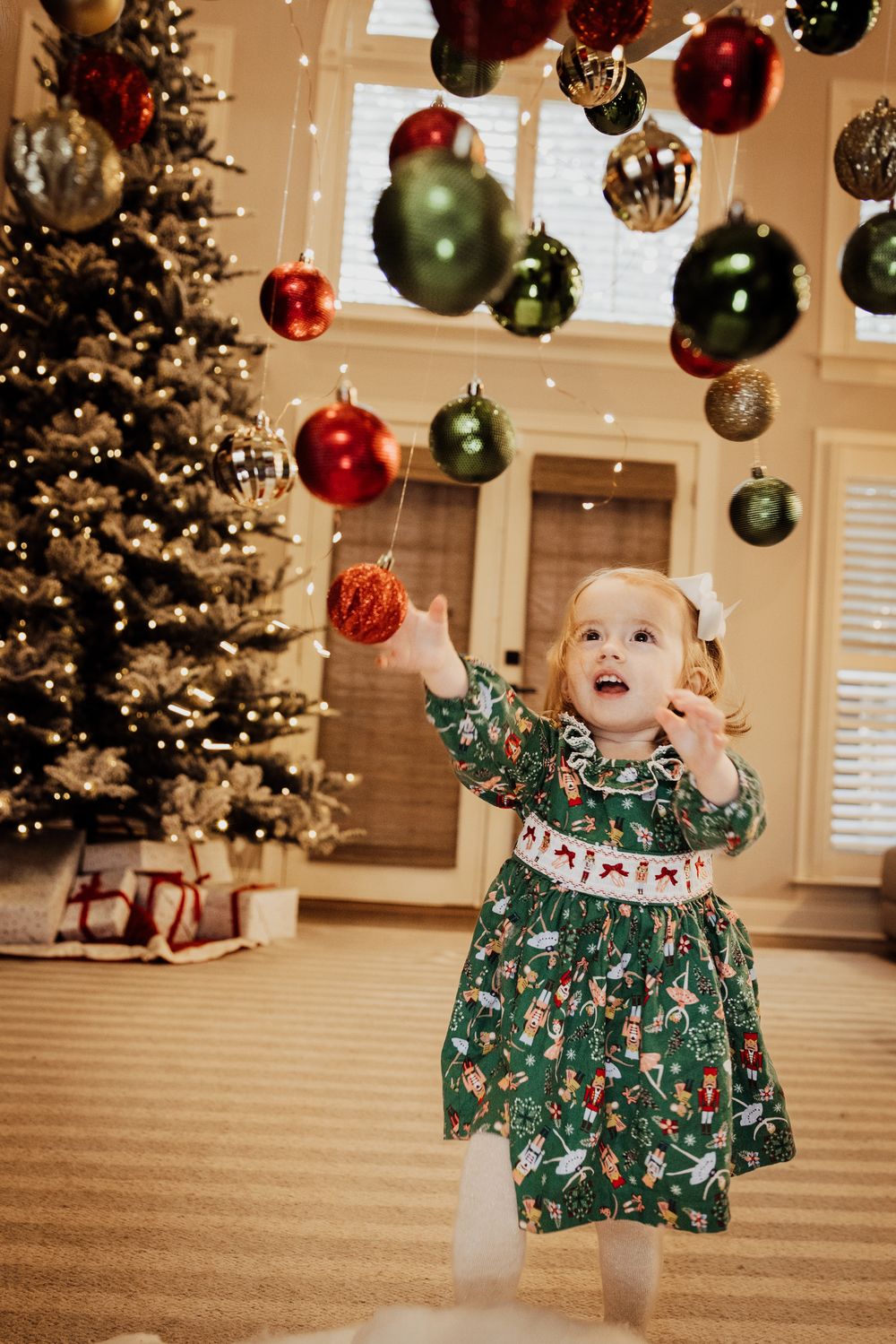 Toddler girl in green Christmas dress reaching for red ornament near decorated holiday tree with hanging baubles.