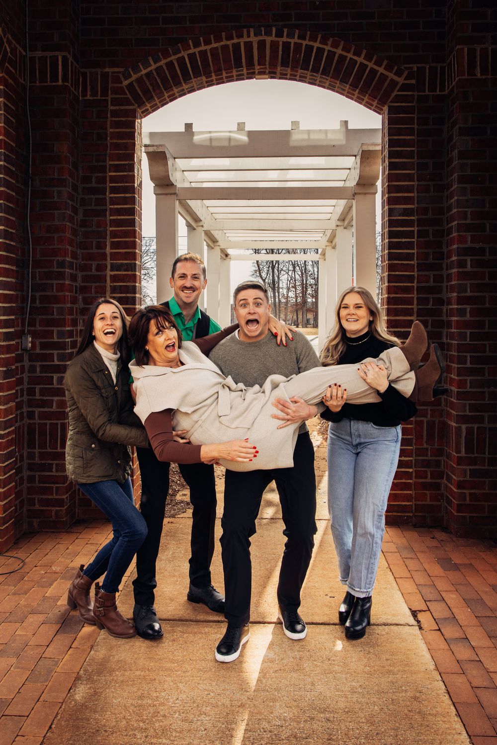 A joyful family of five poses under a brick archway, with one person being carried, all laughing outdoors.