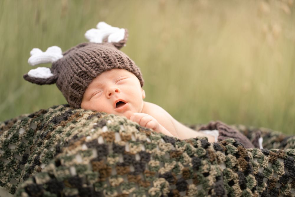 A newborn baby wearing a knit hat yawning while lying on textured fabric outdoors.