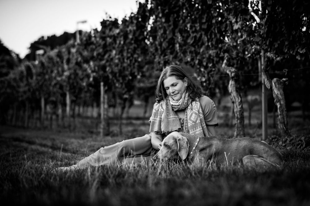 Black and white photo of a smiling woman sitting in a vineyard with a golden retriever dog on a grassy path.