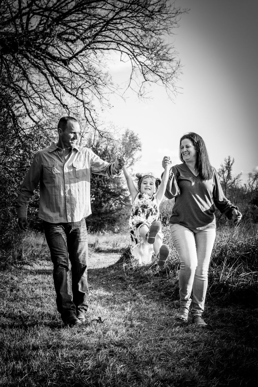 Black and white photo of parents swinging their young child between them while walking outdoors near trees.