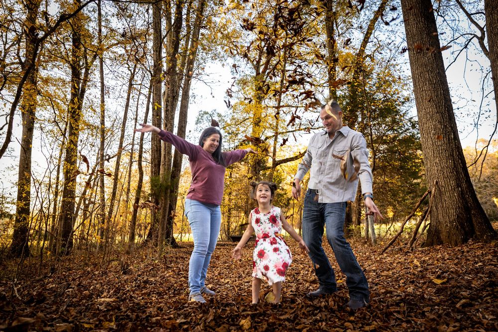 Family playing in autumn leaves among golden trees, parents and young daughter laughing outdoors in fall forest setting.