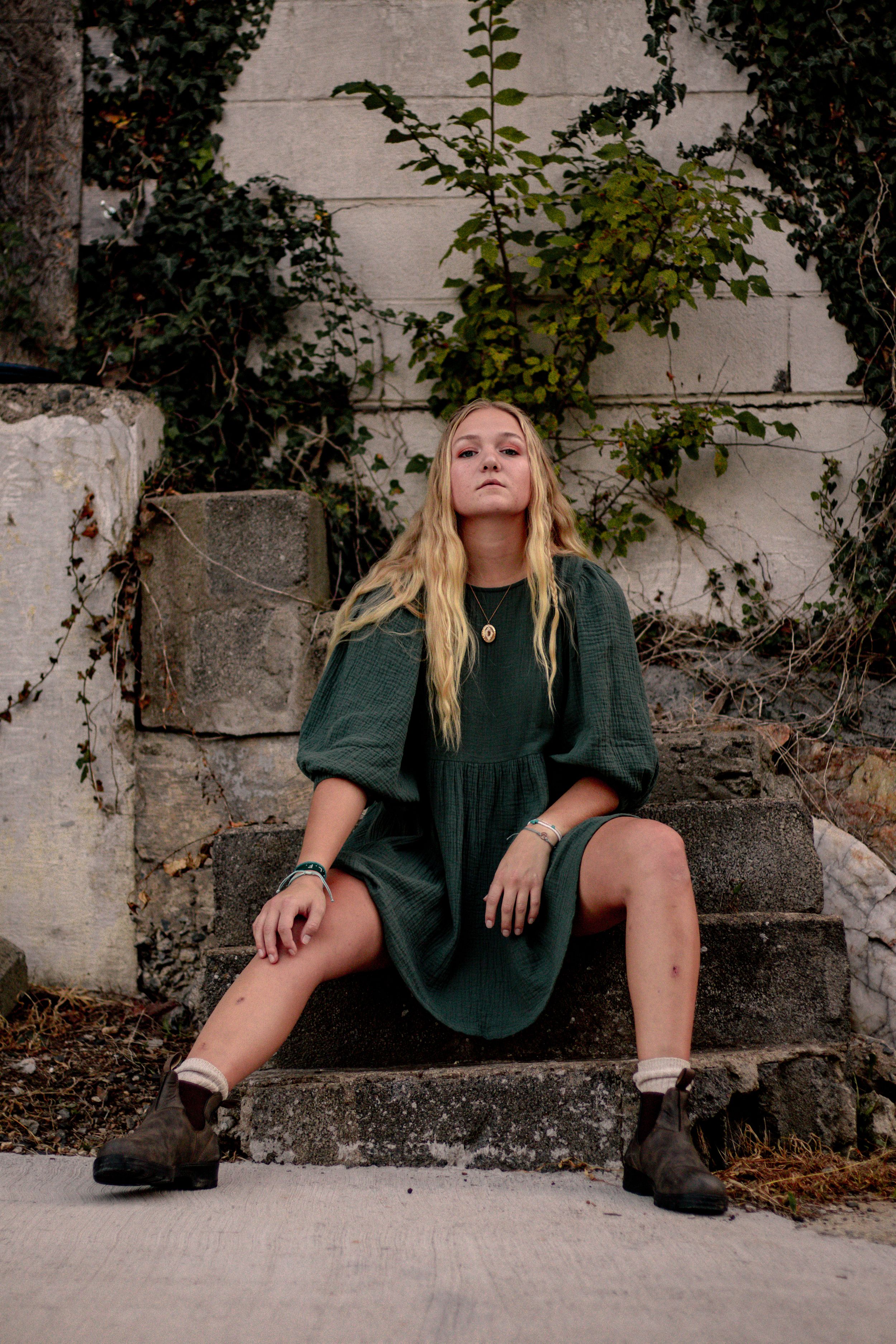 Person in green dress sitting on concrete ledge with urban greenery in background.