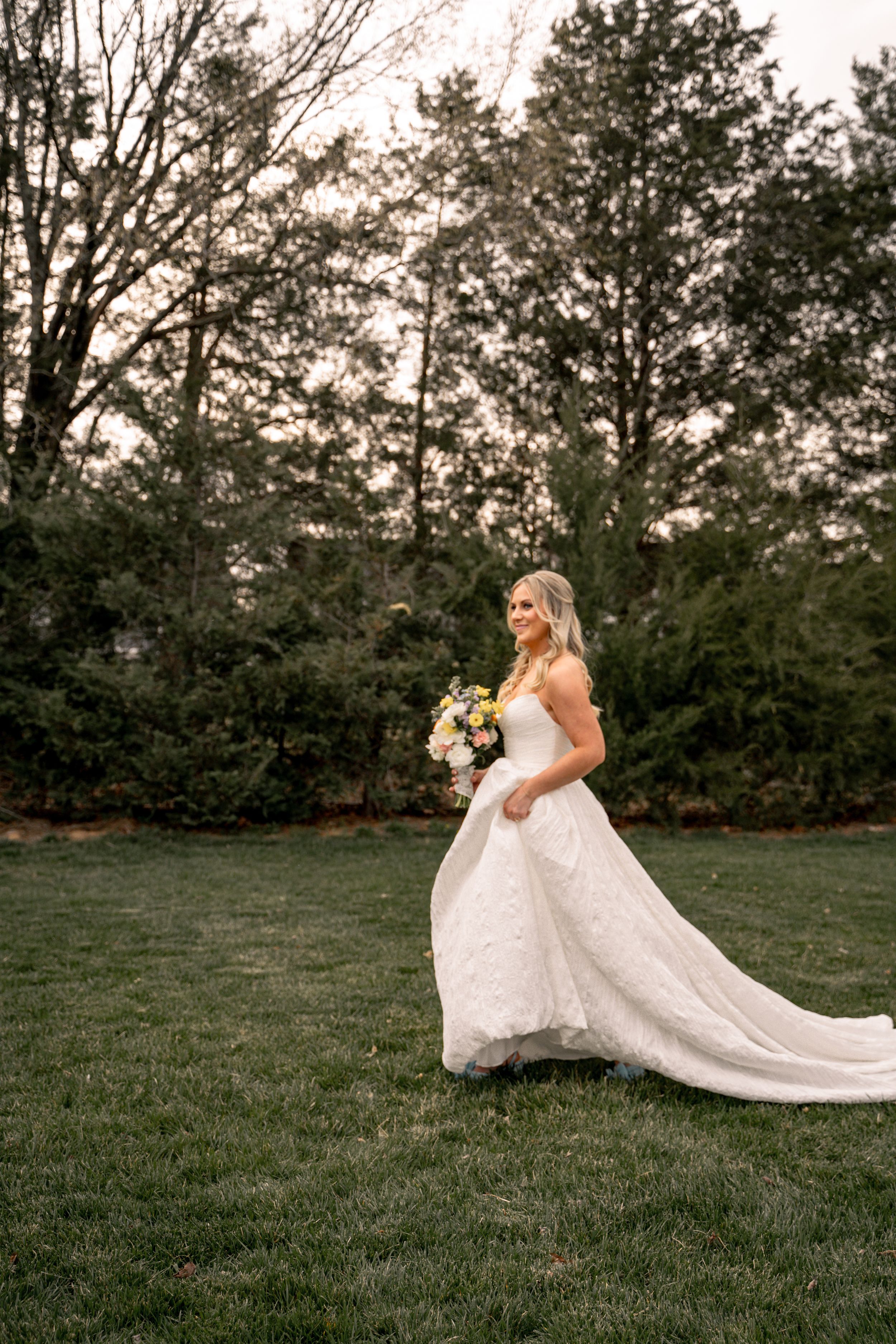 A bride in a flowing white wedding dress sits gracefully on a grassy hill during golden hour.