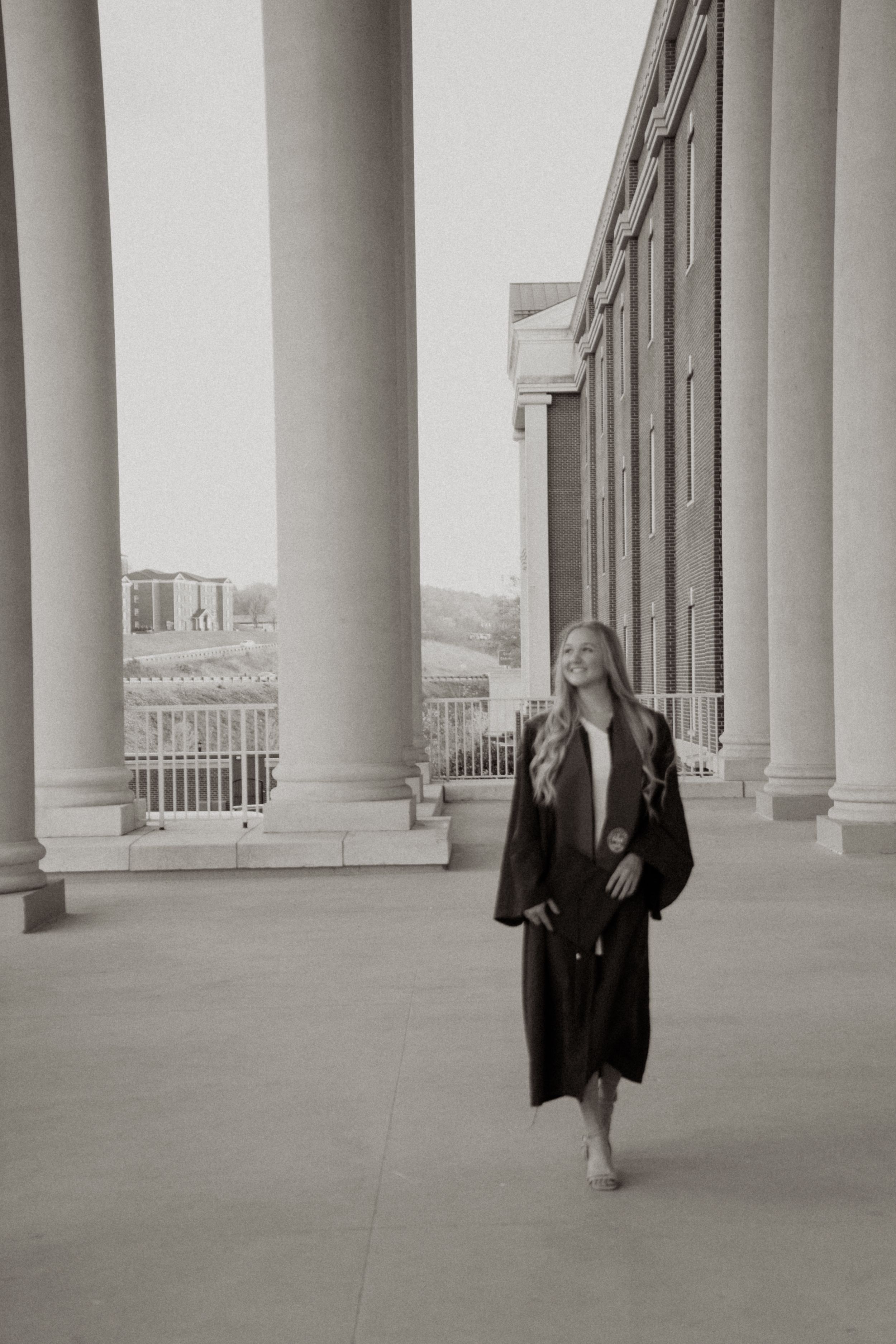 Black and white photo of a person walking between classical columns in a long coat and dress. College graduation.