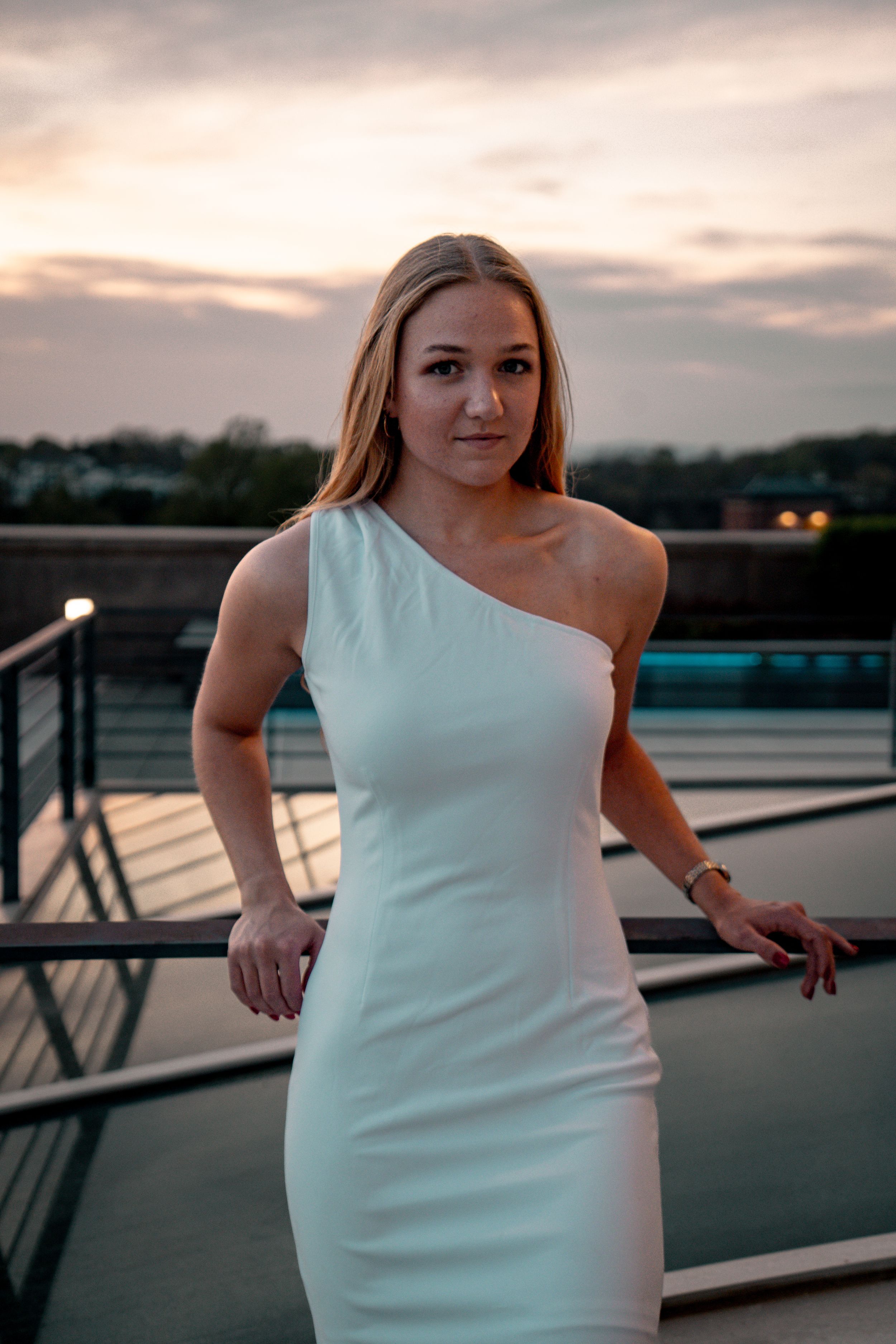 A person in a white formal dress poses against a sunset railing overlooking water during golden hour.
