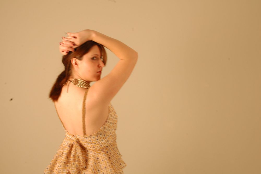 Woman in backless beige dress with gold choker necklace posing against neutral background.