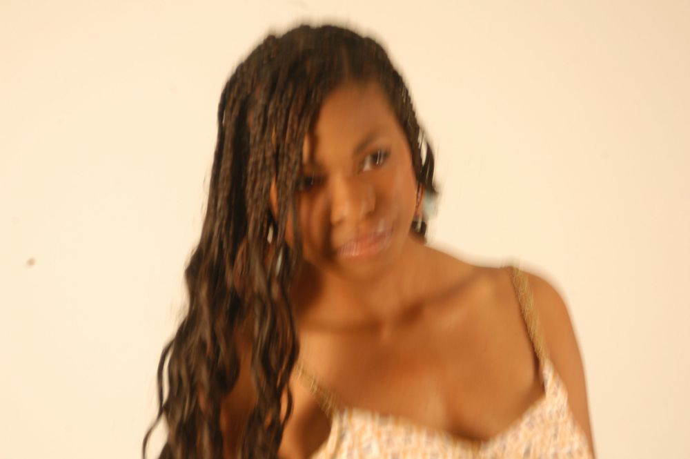 Young Black woman with long braided hair smiling softly in a white top against white background.