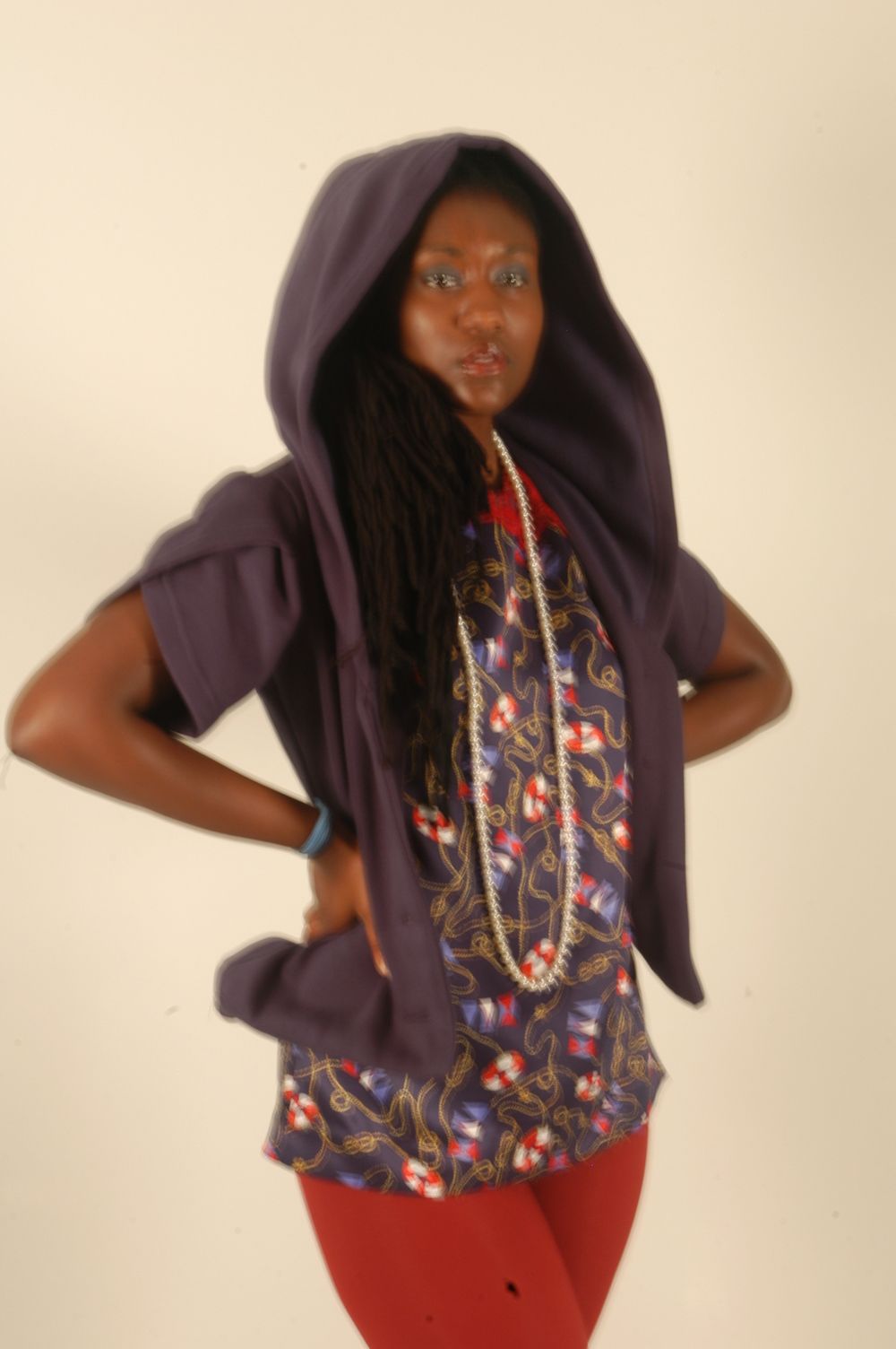 Black woman in purple hooded vest with dreadlocks and red pants posing against white background.