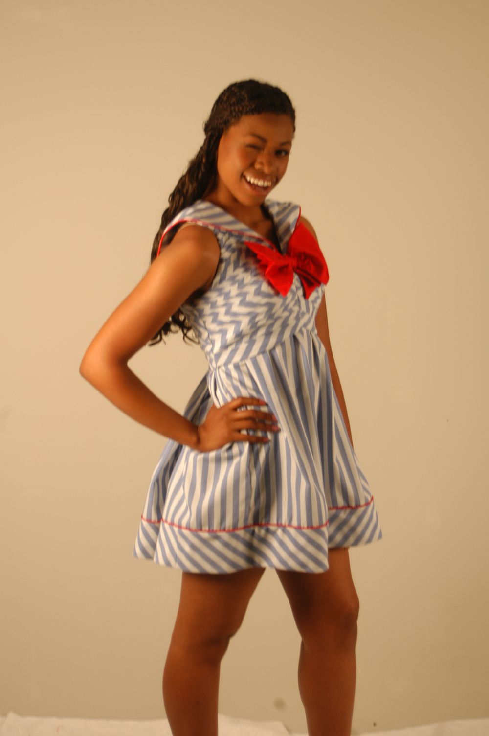 Smiling young woman in blue striped sailor dress with red bow posing against beige background.