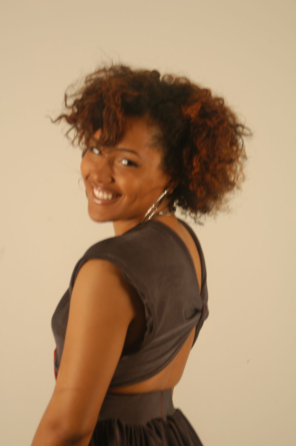 Smiling Black woman with curly ponytail in backless gray dress posing against beige background.