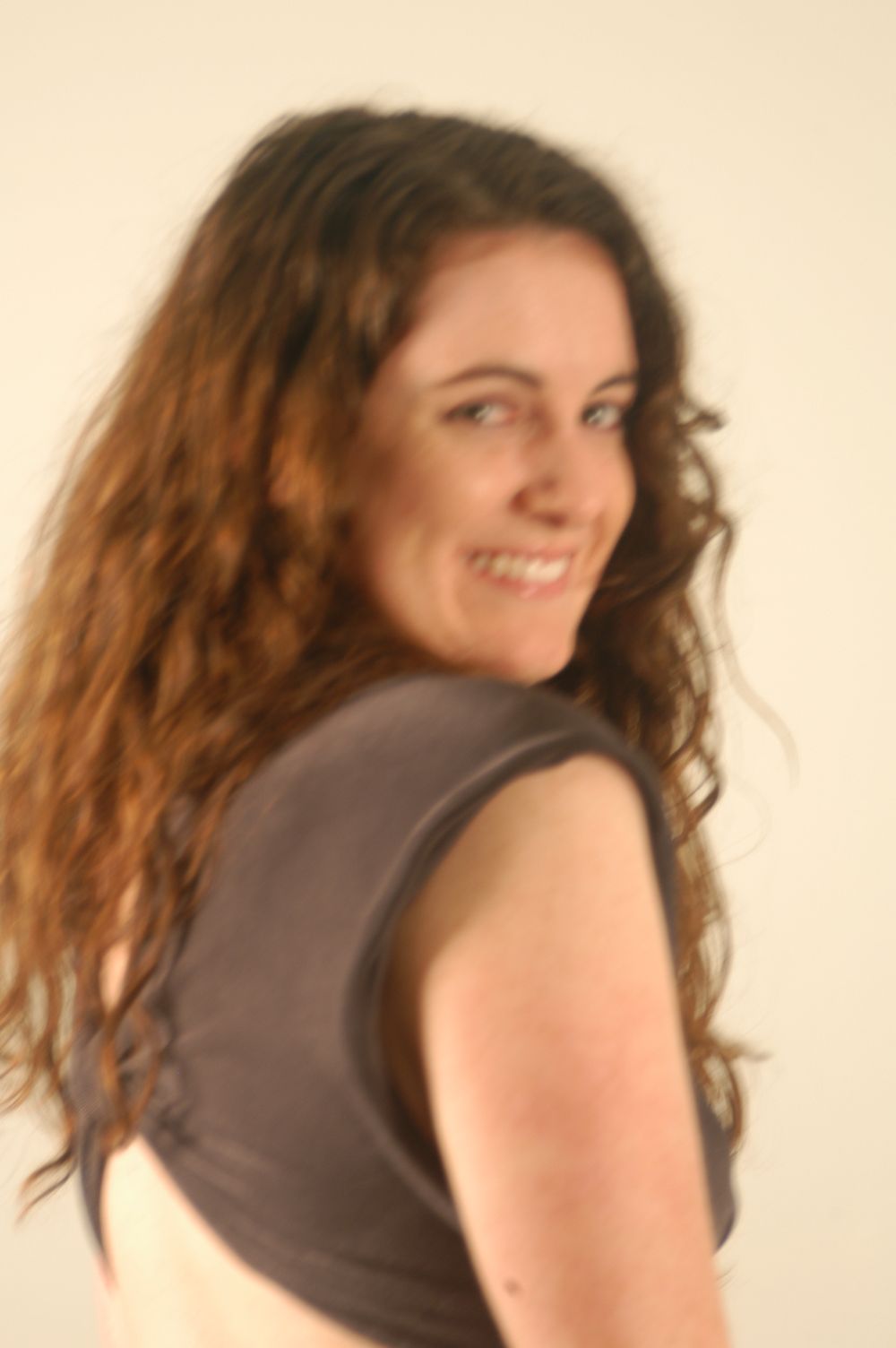 Young woman with long curly brown hair smiling over her shoulder wearing a gray sleeveless top.