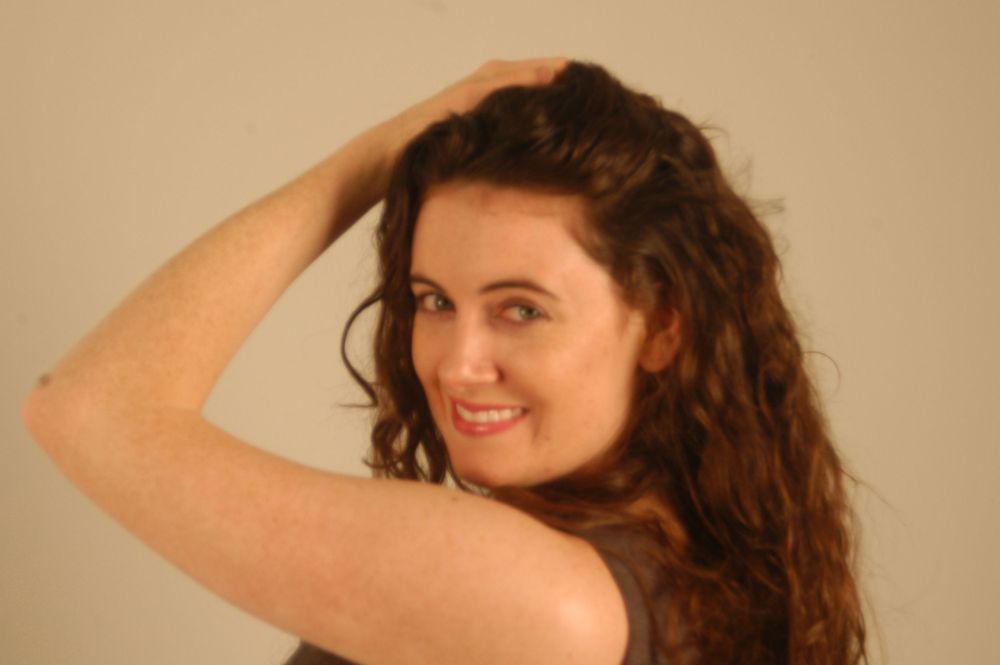 Young woman with long curly brown hair smiling while holding her hair up with one hand.