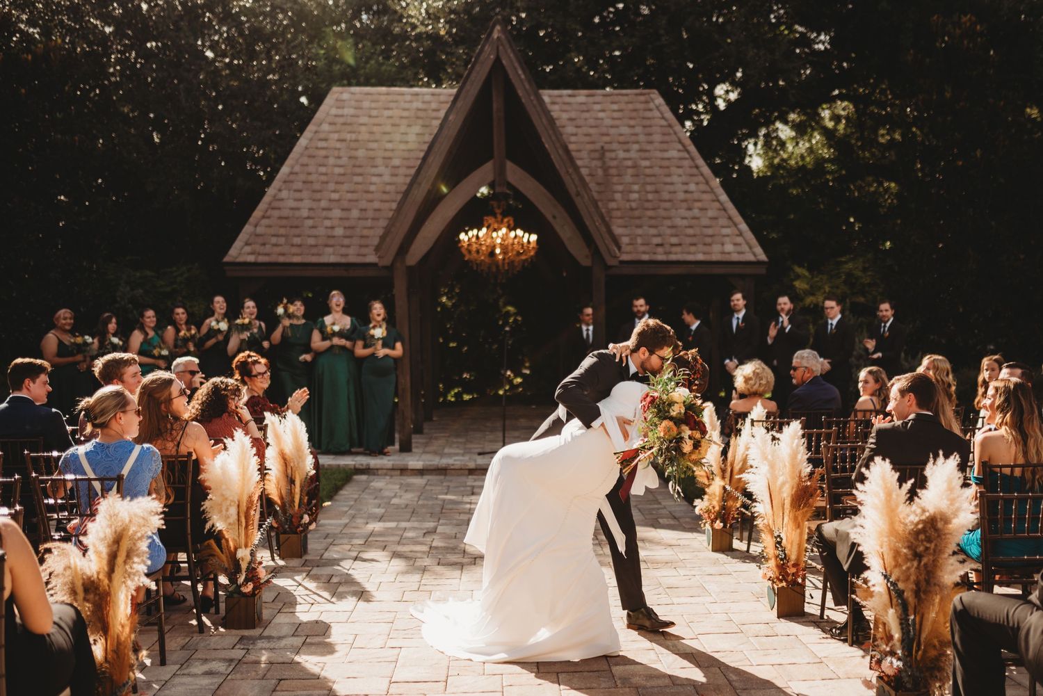 Wedding ceremony taking place under a wooden chapel with pampas grass decorations at sunset.