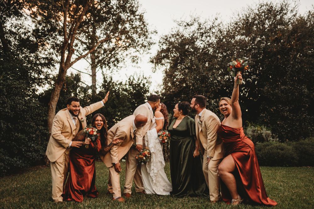 Wedding party in beige and burgundy attire poses playfully on a lawn at sunset.